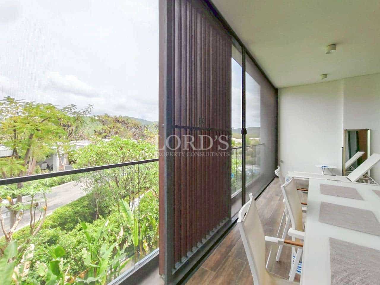 Modern apartment balcony with glass railing, outdoor dining chairs, and lush green view.