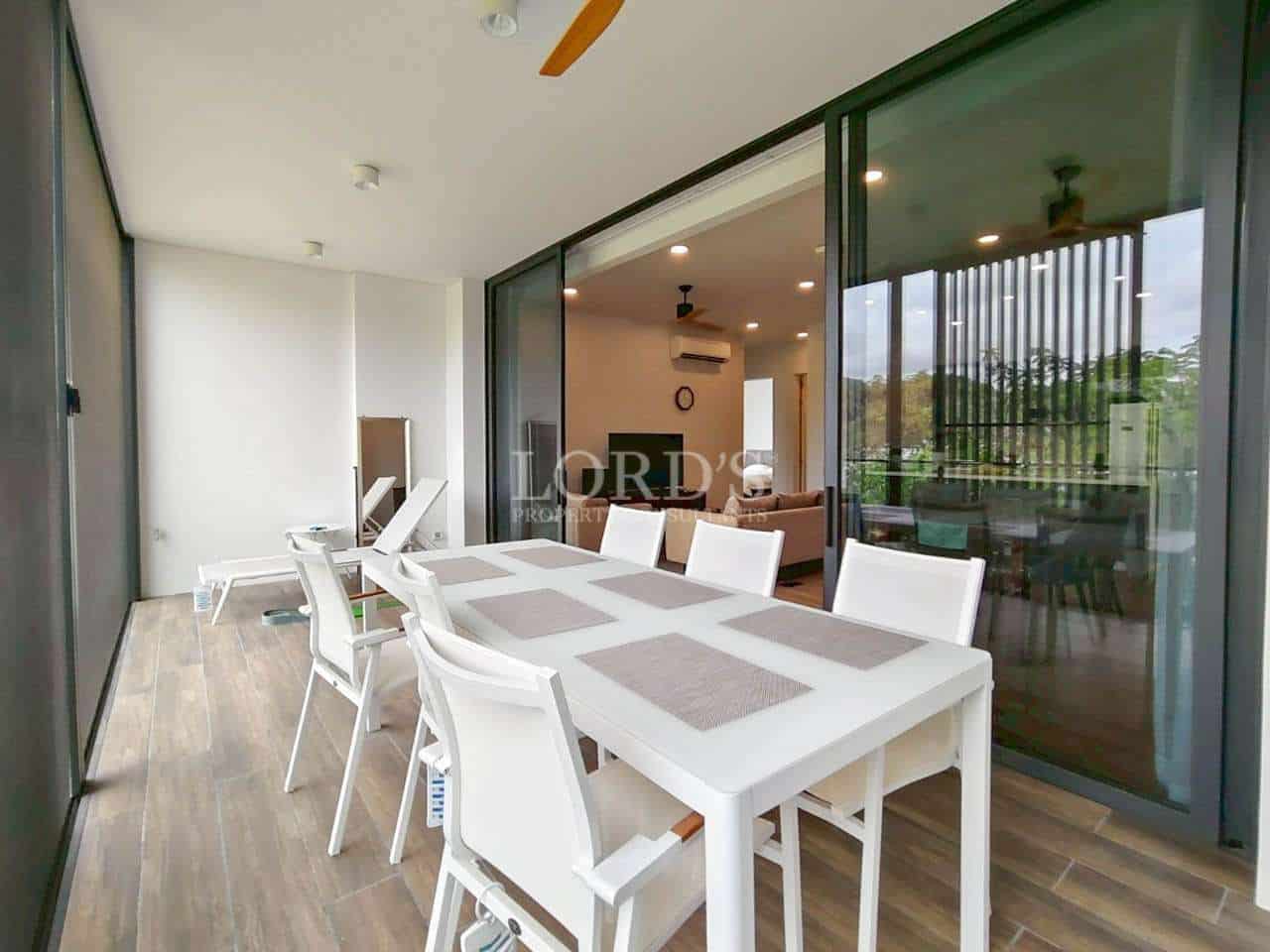 Modern balcony dining area with white table and chairs, sliding glass doors opening to a bright living room.