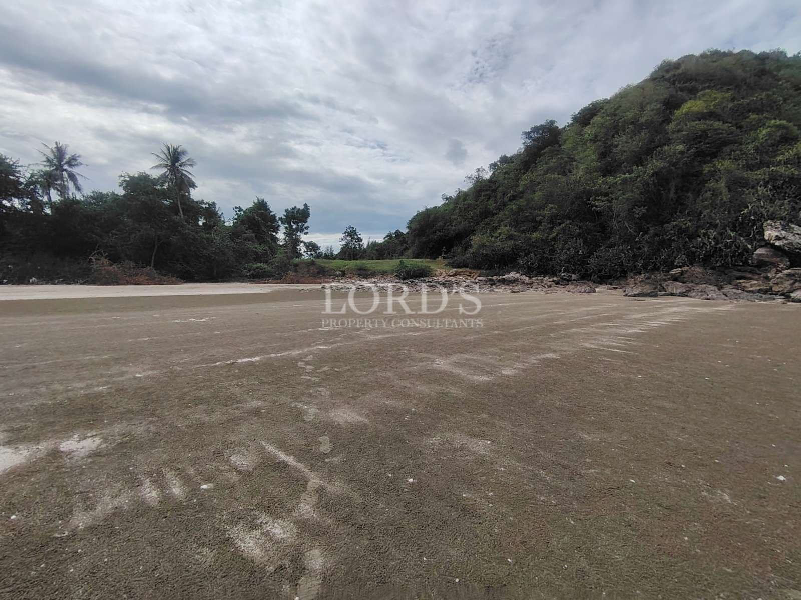 Wide sandy beachfront with calm water, rocky edge, and green hillside under a cloudy sky.