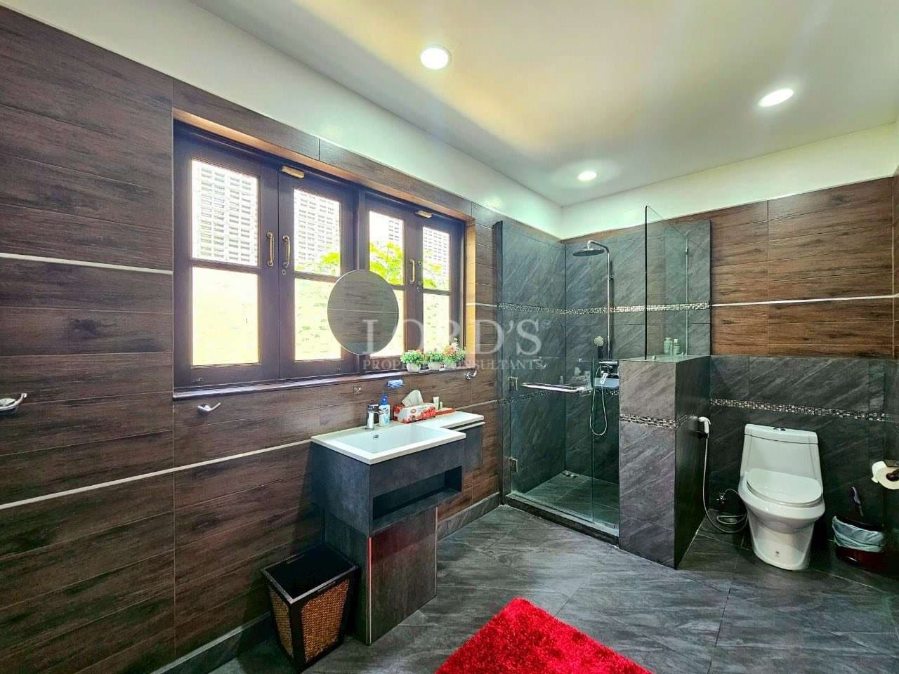 Modern bathroom interior with walk-in glass shower, wooden wall tiles, vanity sink, and natural light.