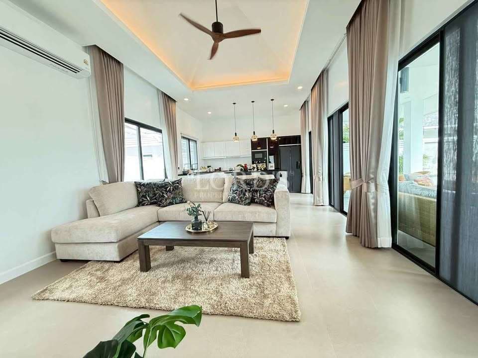 Modern open-plan living room with beige sectional sofa, ceiling fan, large windows, and kitchen in the background.