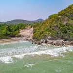 Aerial view of a secluded beach with gentle waves, rocky headland, and lush green hills in the background.