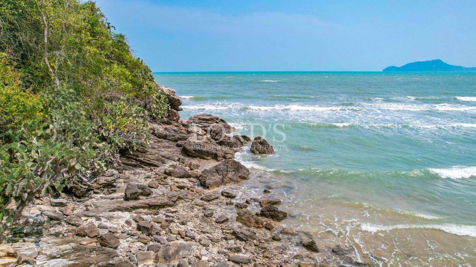 Rocky tropical coastline with green vegetation, gentle ocean waves, and a clear blue sky.