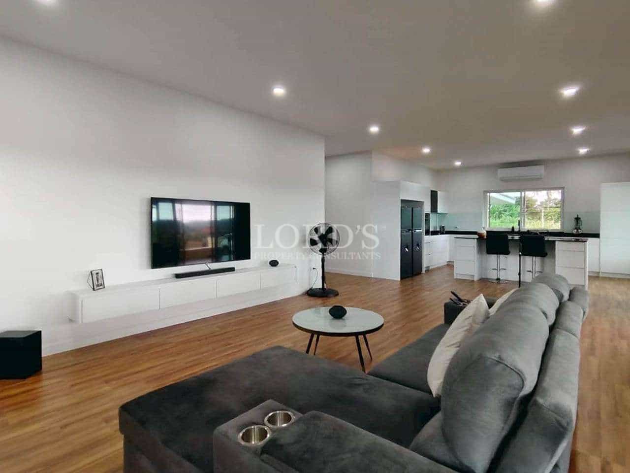 Open-plan living room and kitchen with grey sofa, wall-mounted TV, wooden flooring, and modern lighting.
