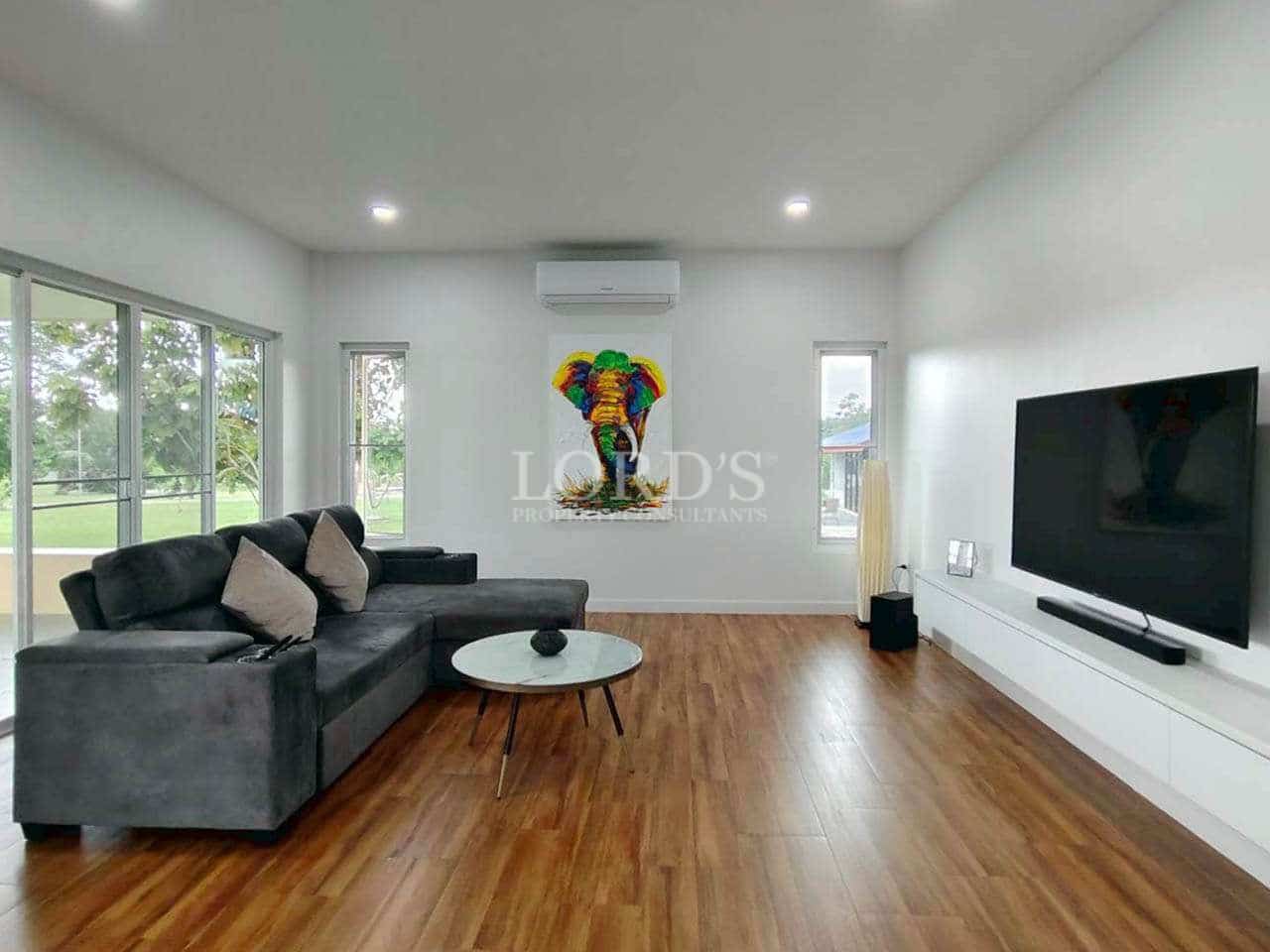 Modern living room with grey sectional sofa, wooden flooring, wall-mounted TV, and large windows overlooking a garden.