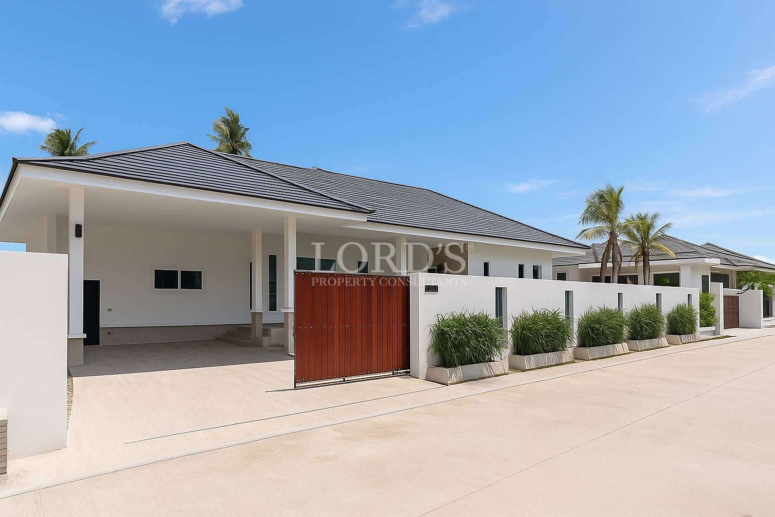 Modern single-story villa exterior with covered carport, wooden gate, white boundary wall, and palm trees under a clear blue sky.