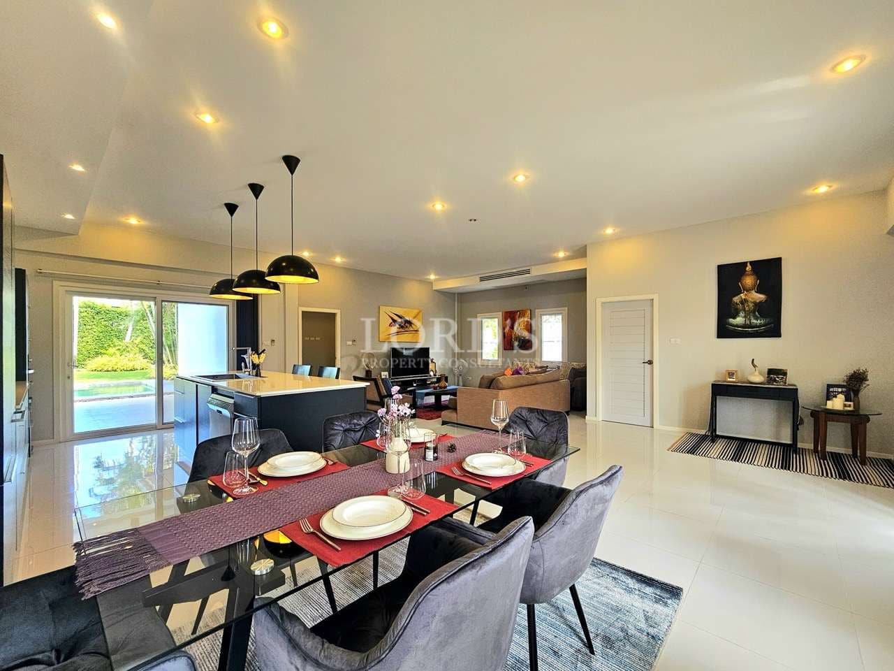Modern open-plan dining and living area with glass dining table, stylish chairs, pendant lighting, and a sleek kitchen island connected to a bright lounge space.