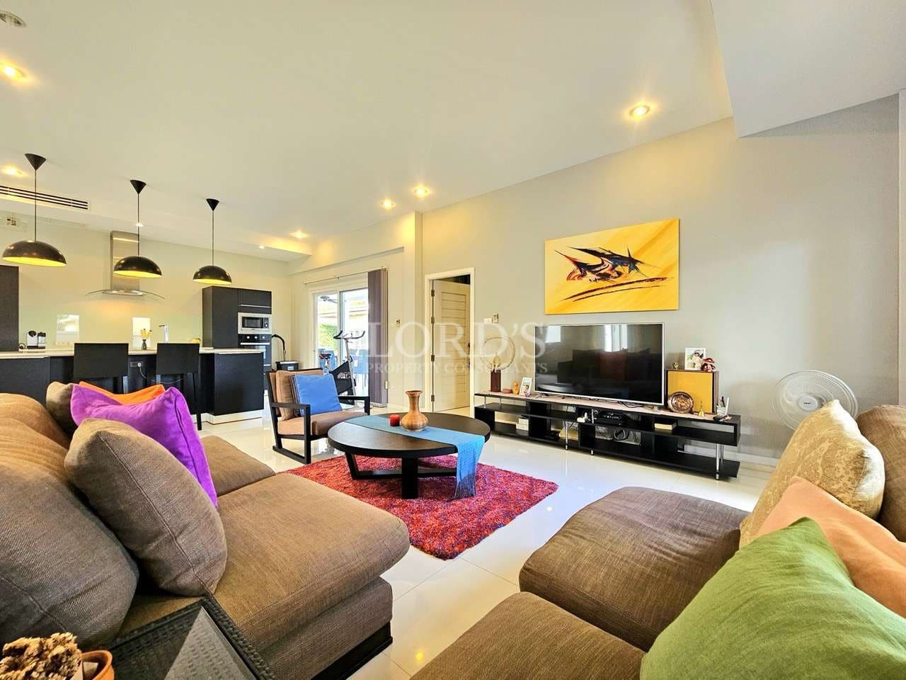 Open-plan living room with modern sofa set, wall-mounted TV, abstract artwork, and a contemporary kitchen in the background.