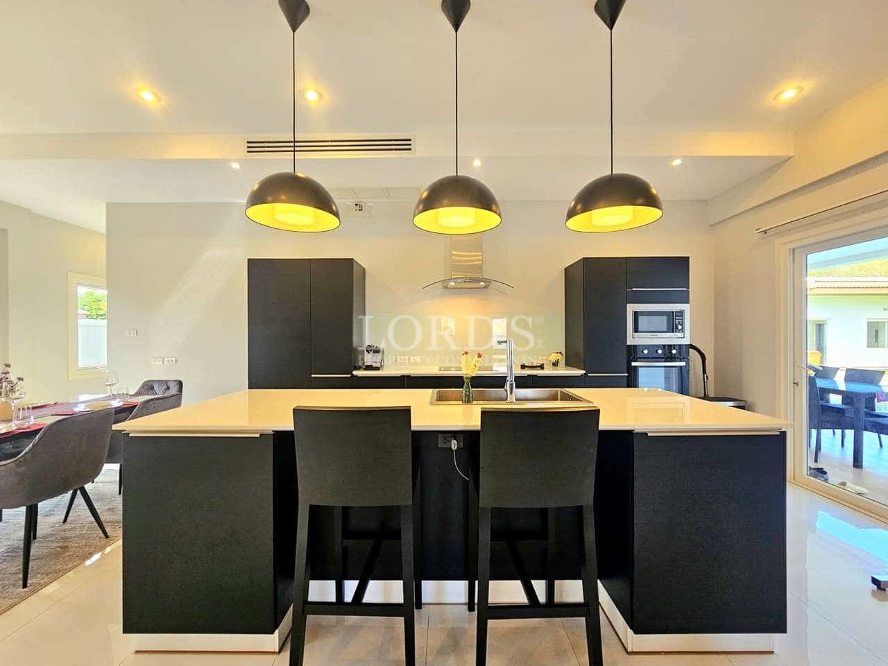 Modern kitchen with large island, black cabinetry, three pendant lights, and built-in appliances in a bright open-plan space.
