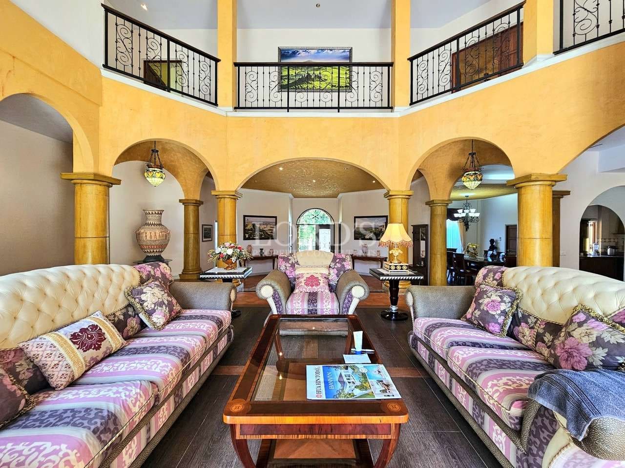 Elegant double-height living room with arched columns, classic sofas, wrought iron balcony railings, and central coffee table.