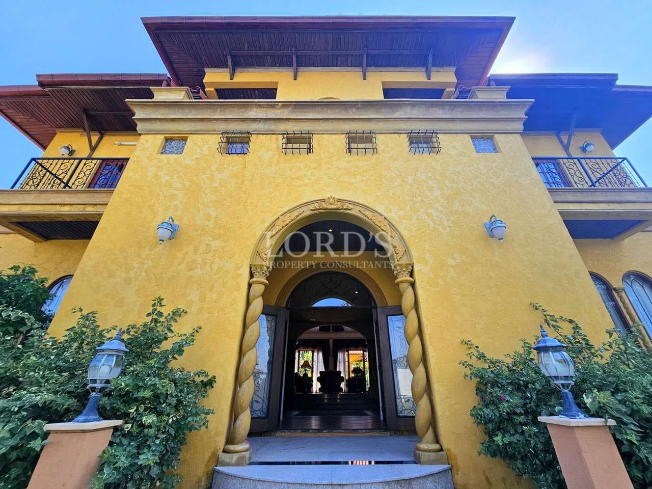Grand Mediterranean-style villa entrance with arched doorway, yellow stucco facade, ornate columns, and upper-level balconies.