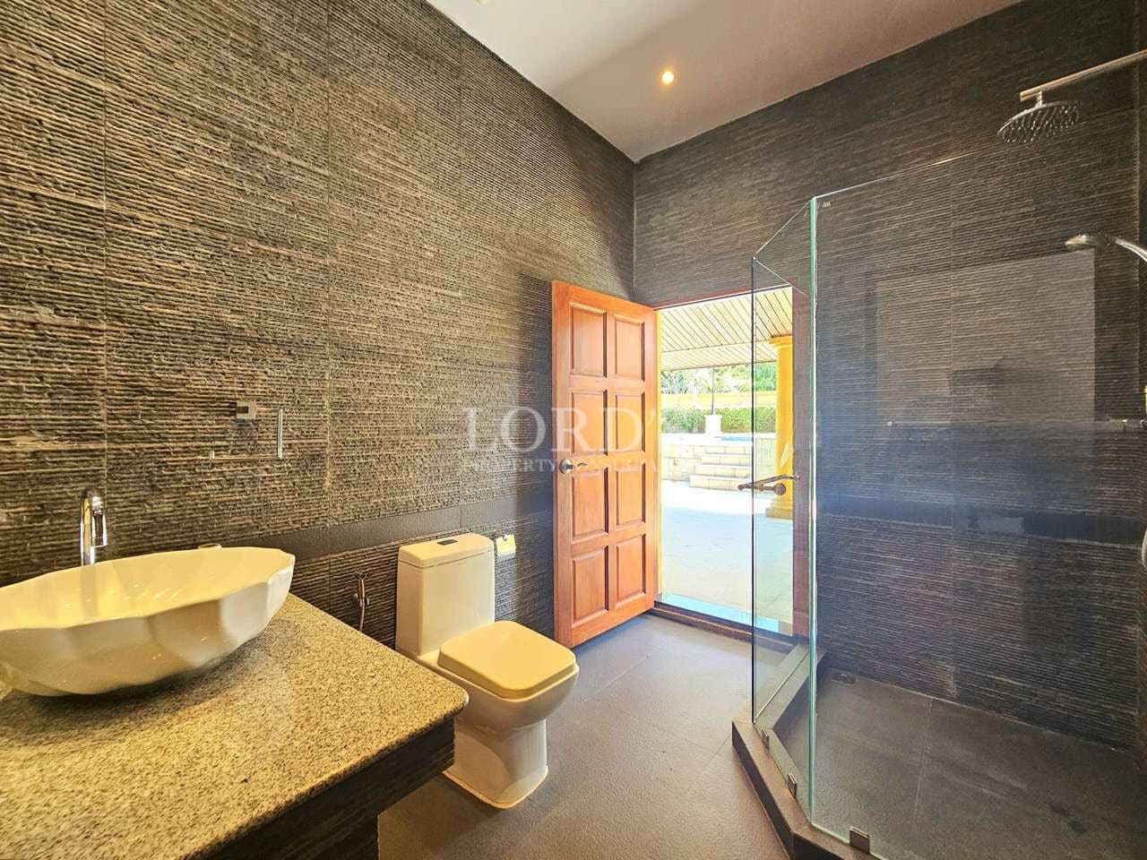Modern bathroom with glass shower enclosure, stone-textured walls, vessel sink vanity, and direct outdoor access through a wooden door.