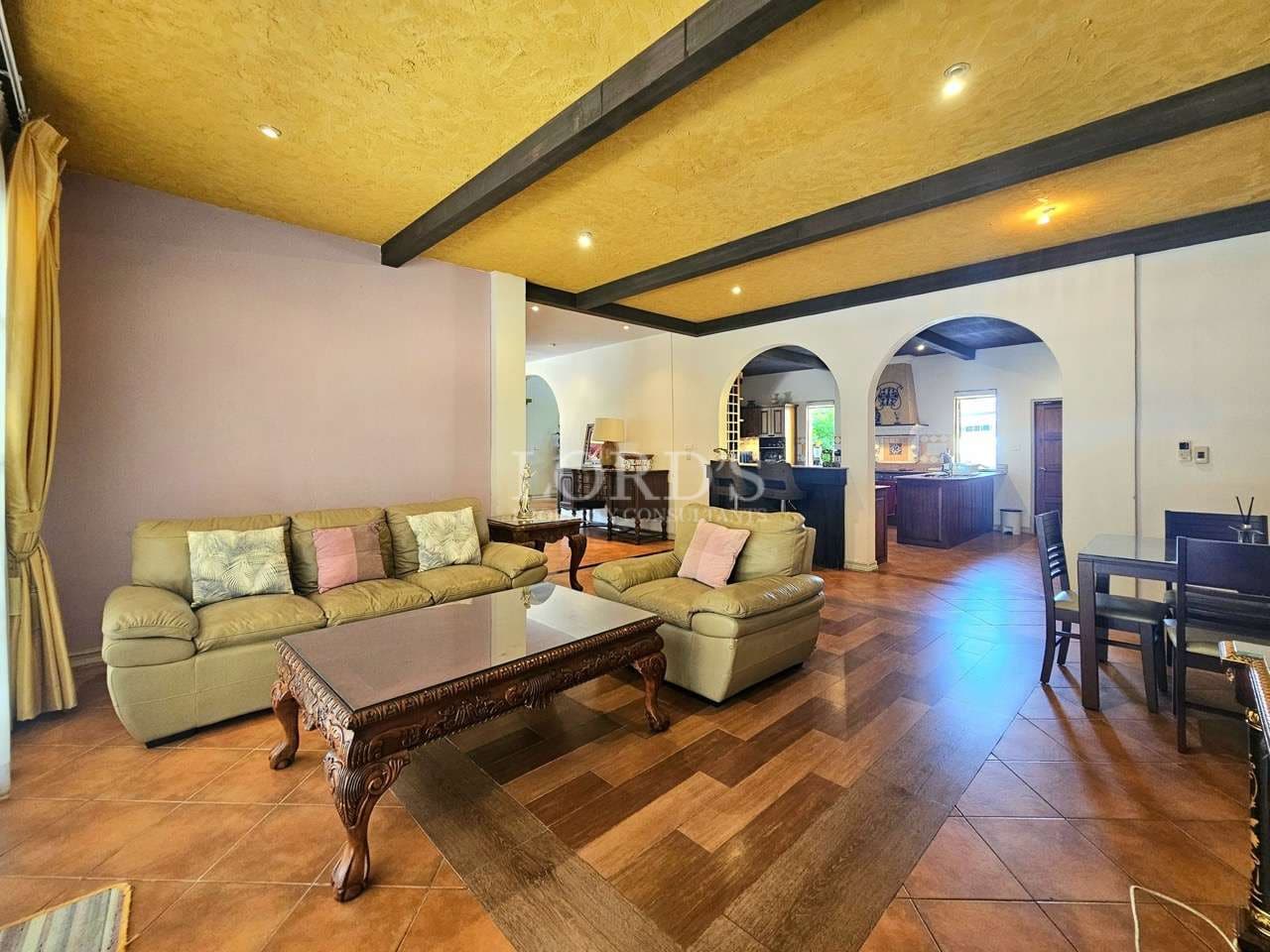 Luxury villa living and dining area with leather sofas, wooden flooring, exposed ceiling beams, and arched open-plan layout.
