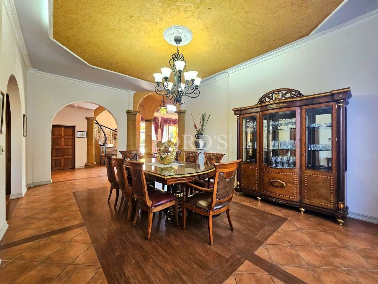 Elegant luxury dining room with classic wooden dining table, chandelier lighting, arched doorways, decorative ceiling, and glass display cabinet.