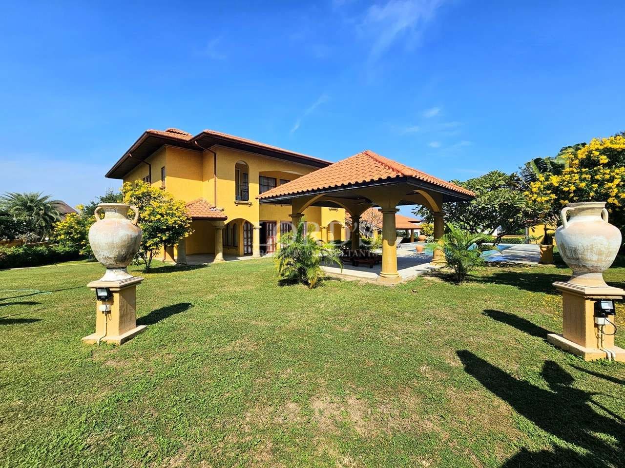 Luxury Mediterranean-style villa with yellow exterior, terracotta roof, private garden pavilion, lush green lawn, and clear blue sky.