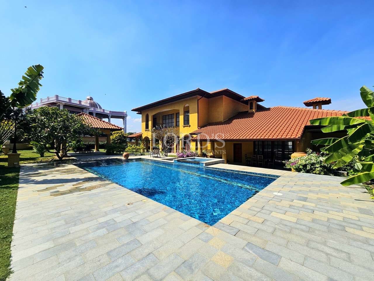 Luxury Mediterranean-style villa with a large private swimming pool, yellow exterior, terracotta roof tiles, landscaped garden, and clear blue sky.