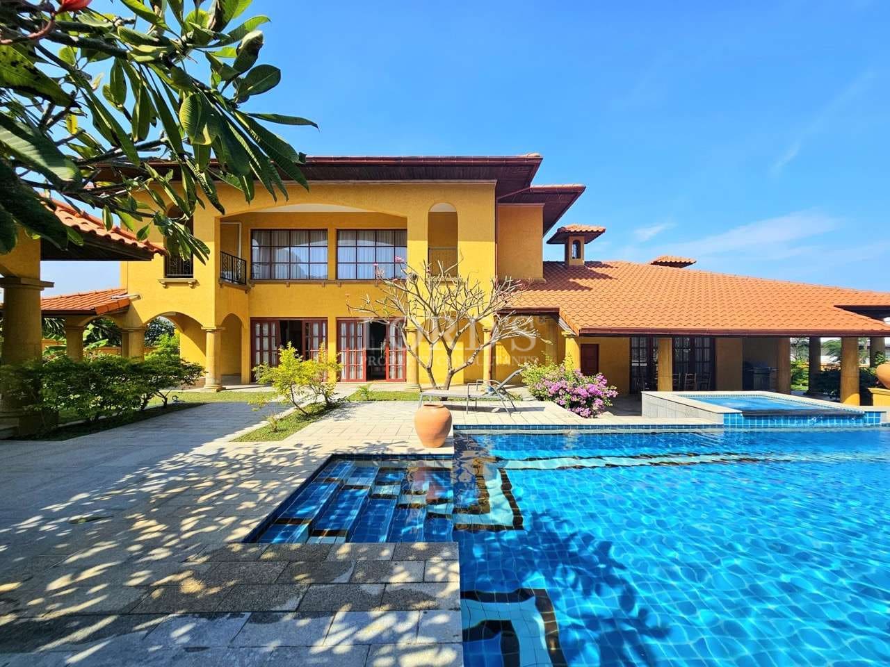 Luxury Mediterranean-style villa with a private swimming pool, yellow exterior walls, tiled roof, landscaped courtyard, and clear blue sky.