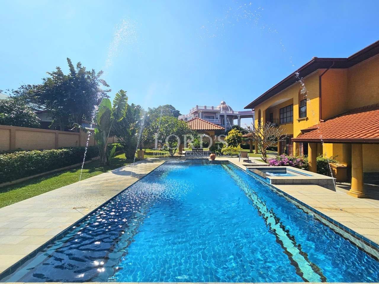 Luxury villa swimming pool with clear blue water, decorative water fountains, landscaped garden, and Mediterranean-style yellow villas under a bright blue sky.
