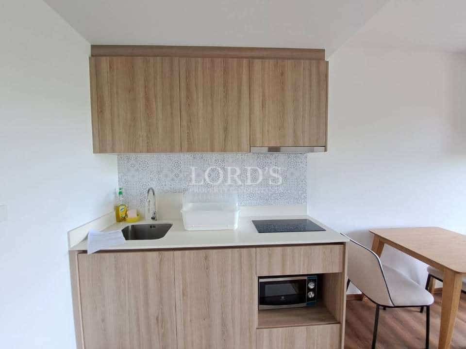 Minimalist apartment kitchenette with wooden cabinets, integrated sink, electric cooktop, microwave storage, and compact dining table.