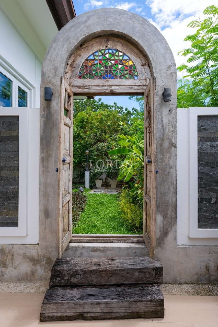 Arched rustic wooden garden gate with stained glass detail opening to a private tropical courtyard.