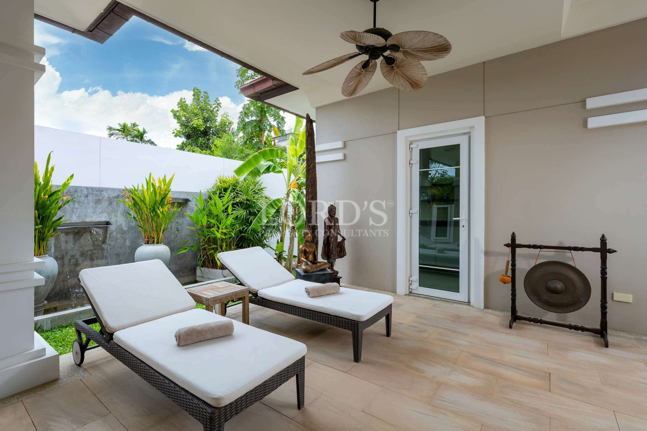 Covered outdoor patio with sun loungers, tropical plants, ceiling fan, and spa-style relaxation area in a luxury villa.
