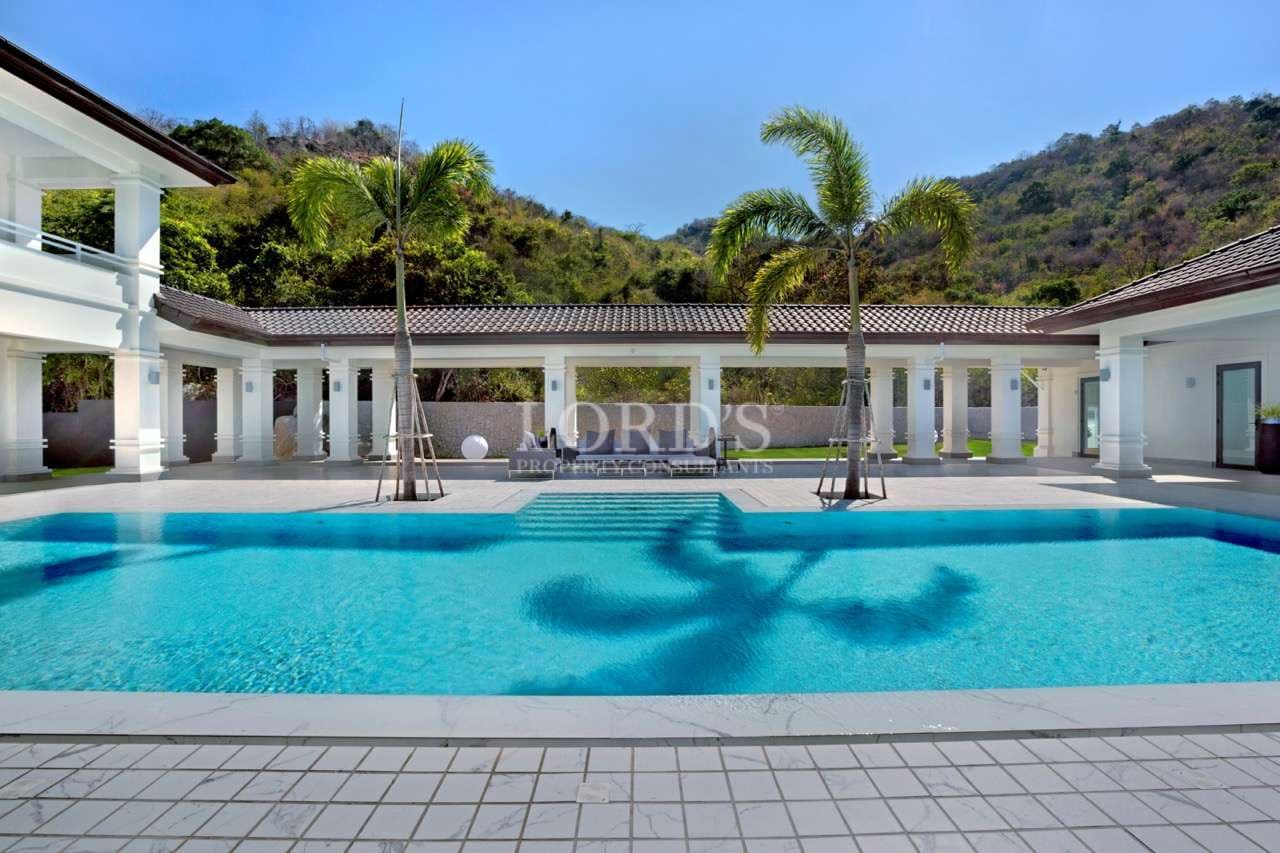 Luxury villa swimming pool with palm trees, white colonnade, and mountain backdrop.