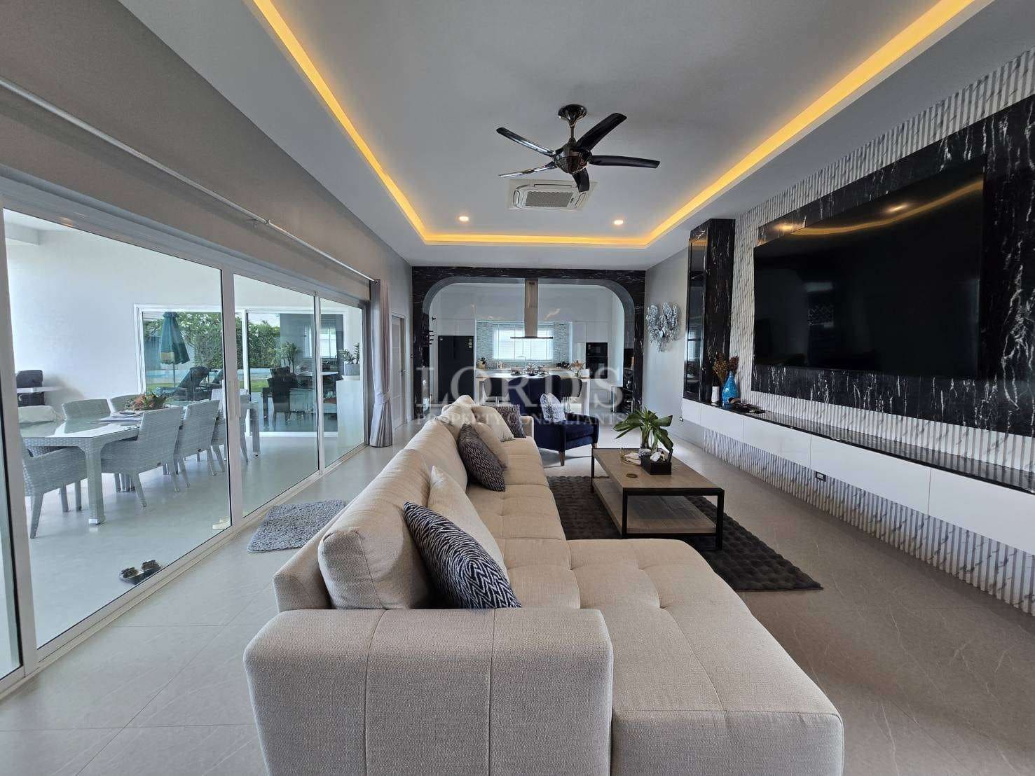 Luxury modern living room with sectional sofa, large wall-mounted TV, LED ceiling lighting, and sliding glass doors.