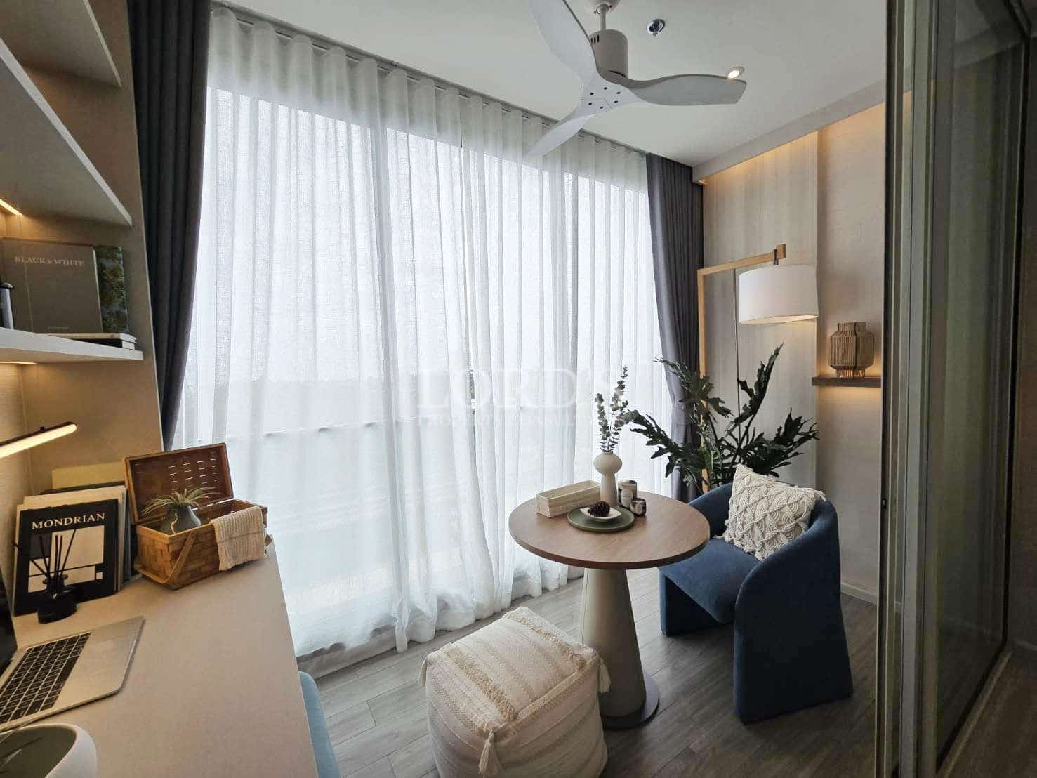 Modern bedroom reading nook with round table, upholstered chair, sheer curtains, ceiling fan, indoor plants, and soft ambient lighting near a window.