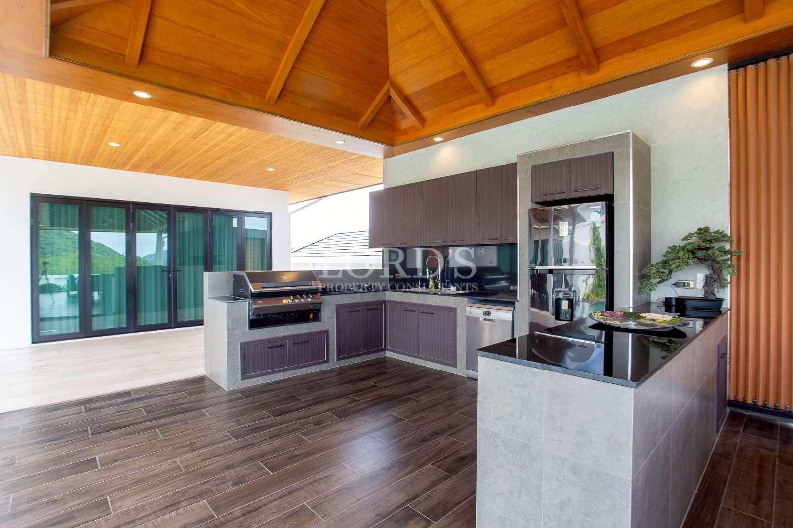 Covered outdoor kitchen with built-in grill, modern cabinetry, and wooden ceiling.
