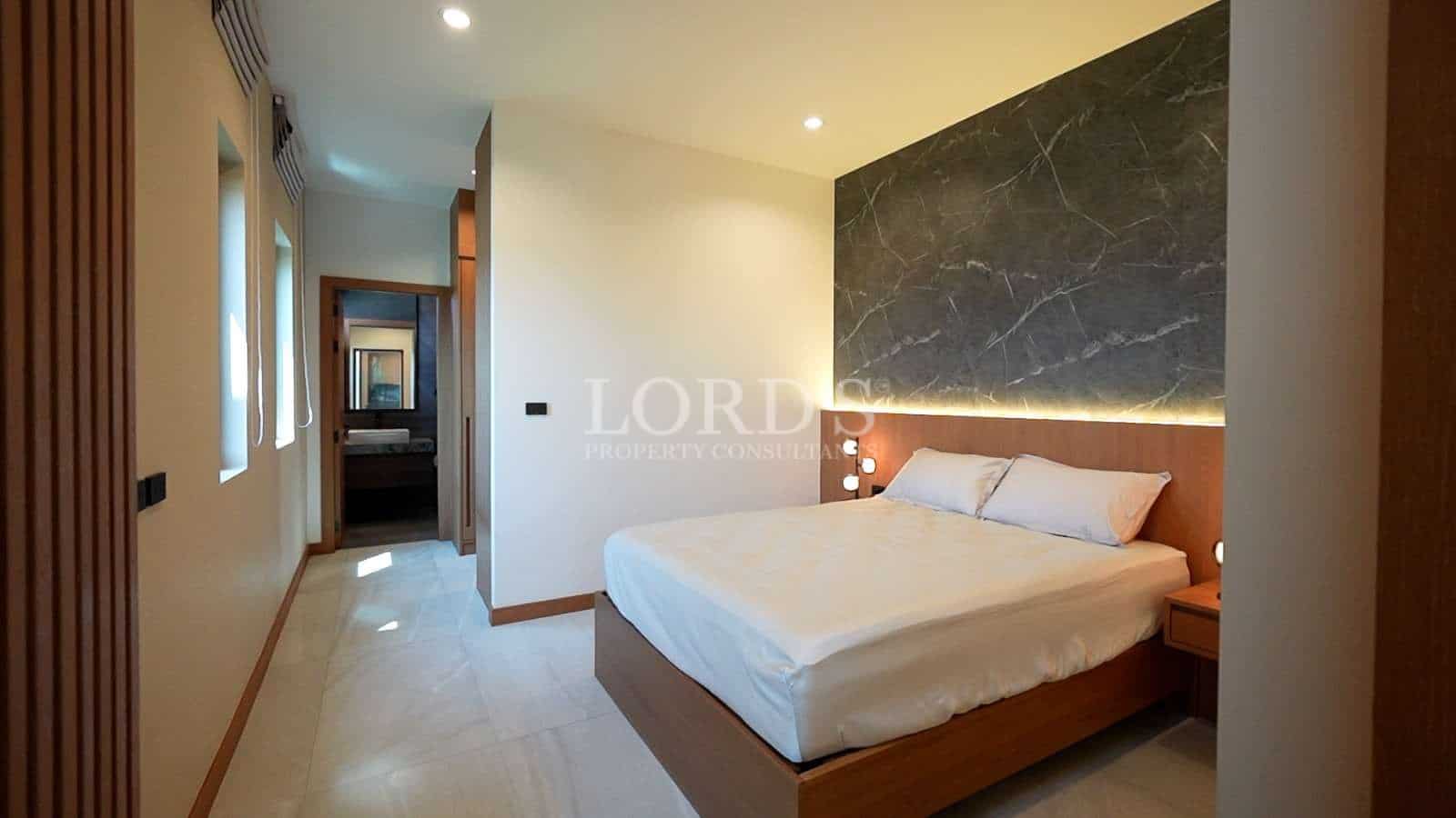 Modern minimalist bedroom with wooden bed frame, marble accent wall, soft lighting, and ensuite bathroom access.