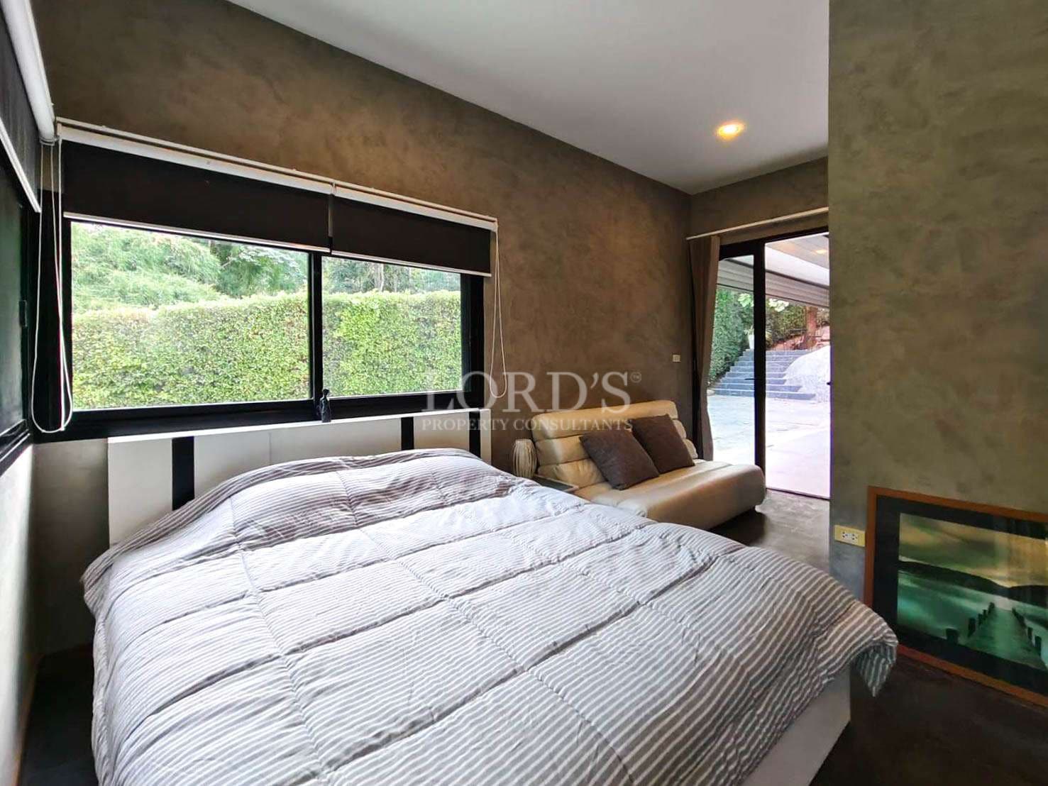 Modern bedroom with large window, striped bedding, sofa seating, and garden view.