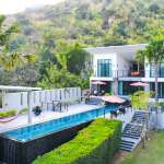 Luxury modern villa with an infinity swimming pool, outdoor terraces, and palm trees set against a lush hillside backdrop.