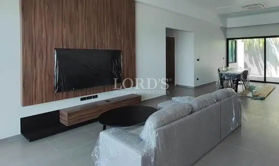 Modern living room with wall-mounted TV, wooden accent panel, sofa seating, and open dining area.