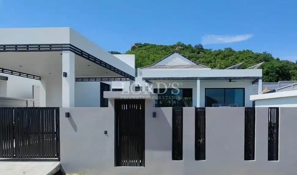 Modern single-story house exterior with flat roof, gated entrance, and hillside background.