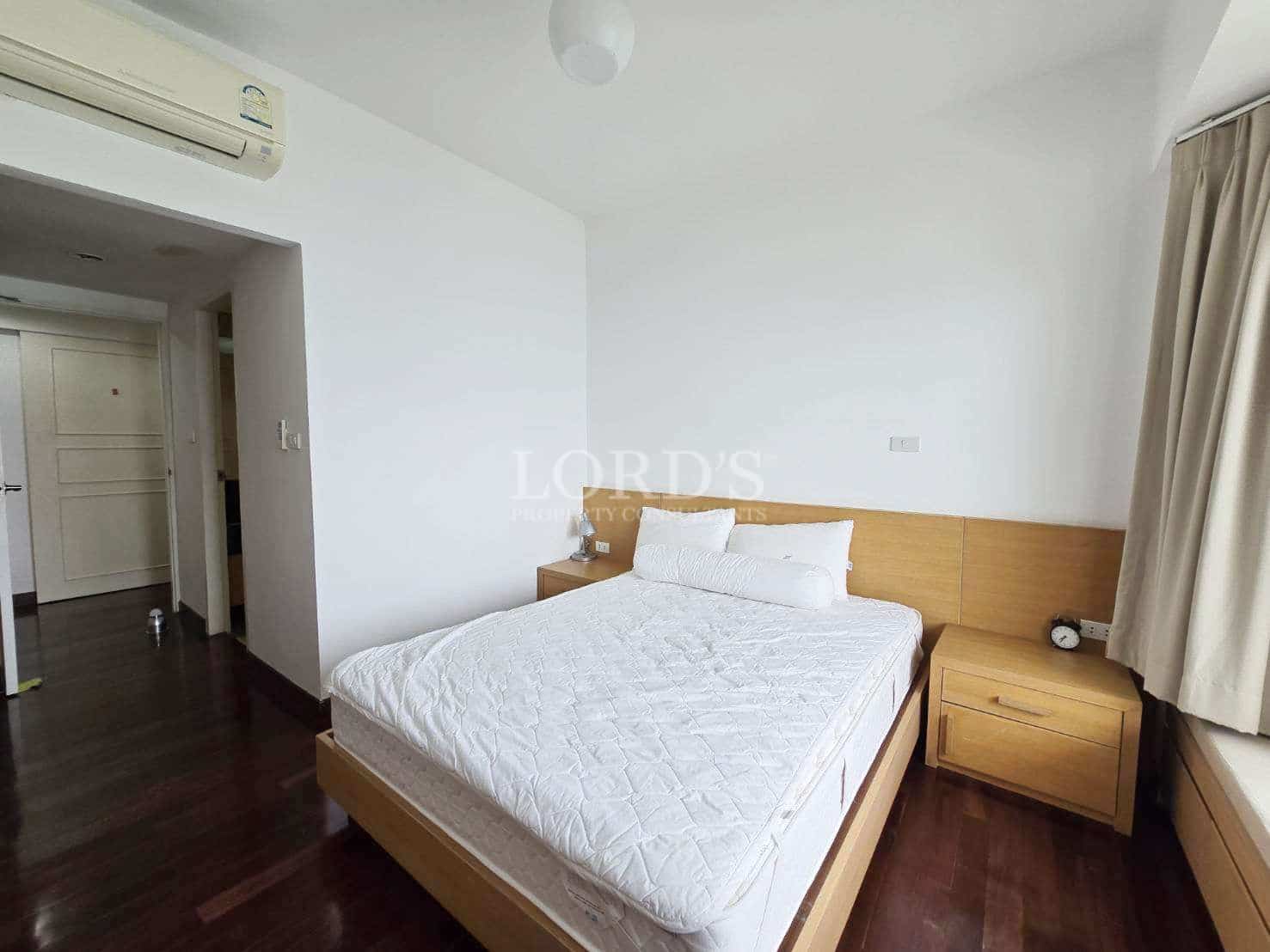 Bright minimalist bedroom with wooden bed frame, white bedding, bedside tables, and air conditioning.
