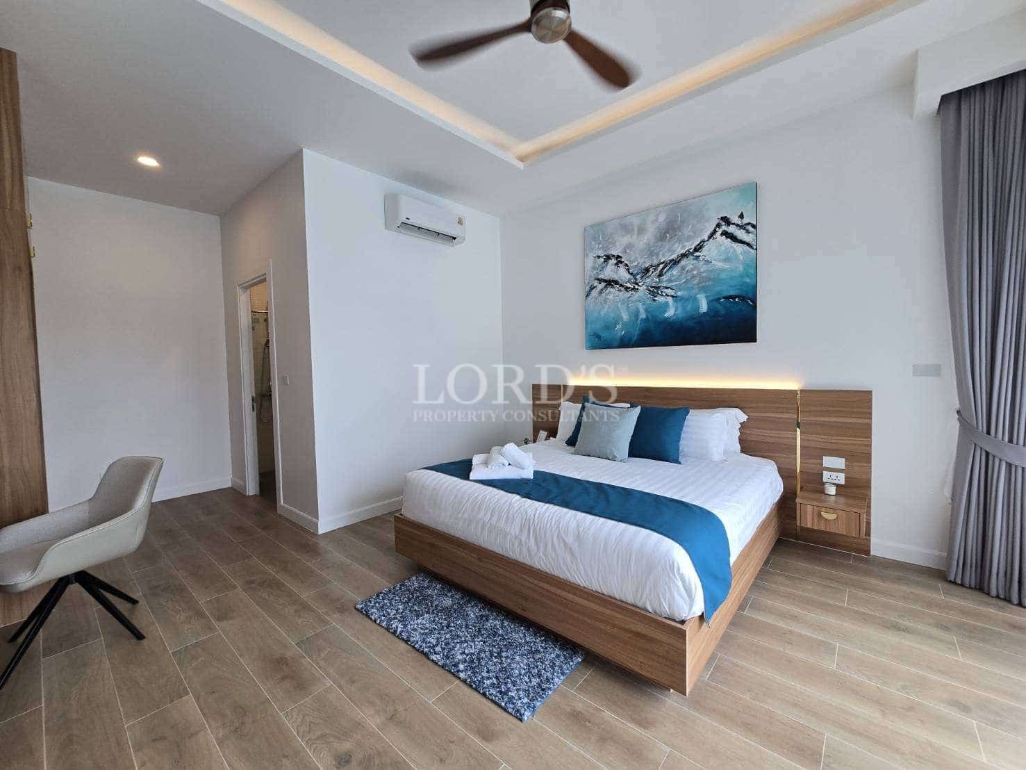 Spacious modern bedroom with wooden bed frame, blue accent bedding, ceiling fan, air conditioning, and wood-look flooring.