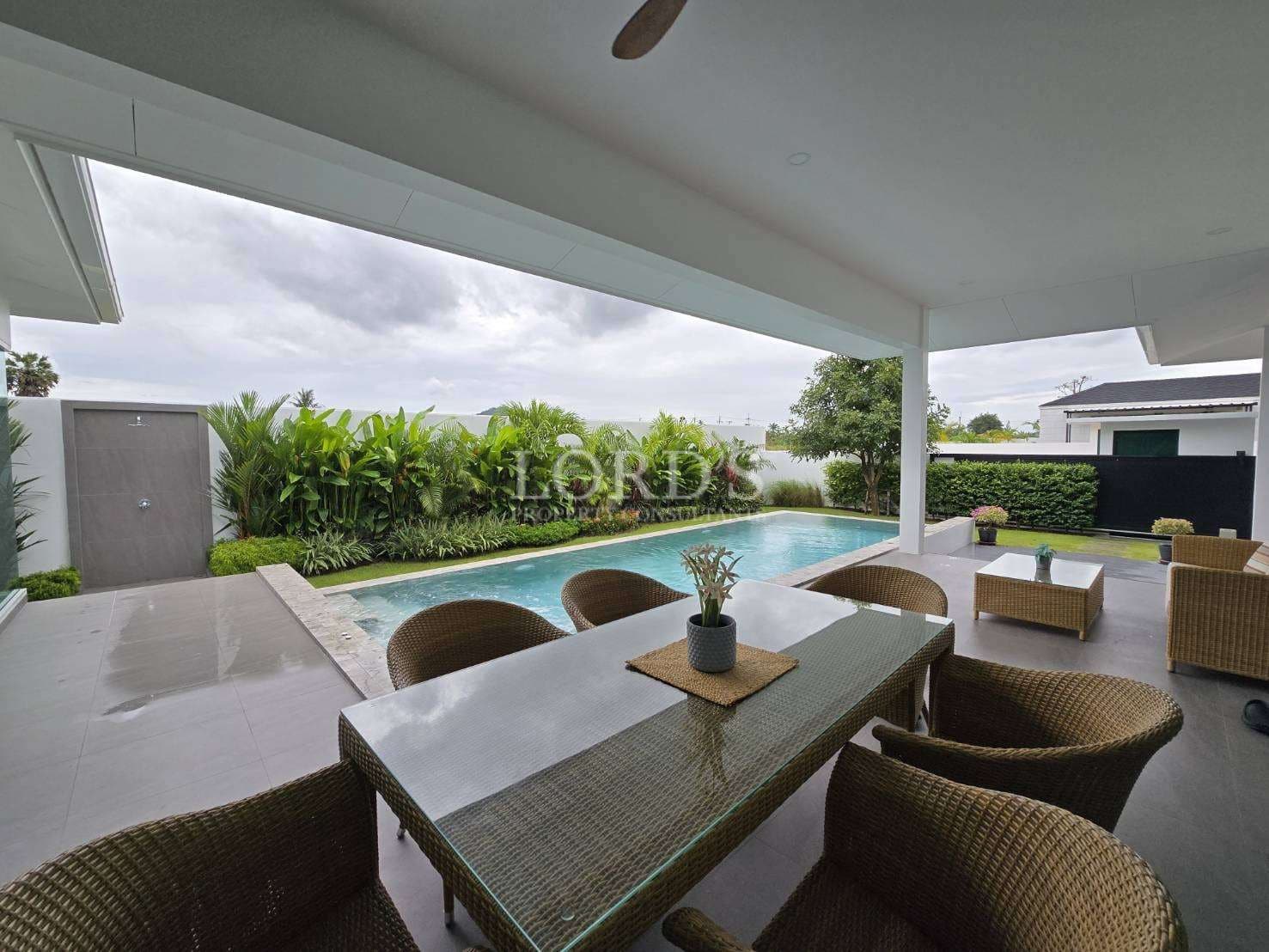 Covered outdoor dining area with wicker furniture overlooking a private swimming pool and garden.