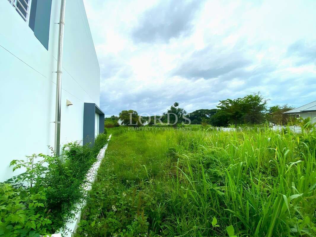 Side view of a modern building with open green land and natural vegetation under a cloudy sky.