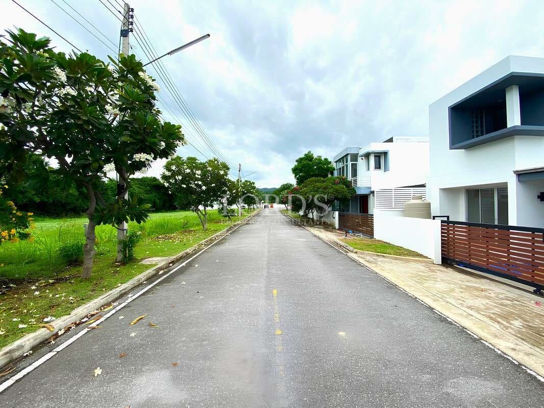 Residential street with modern houses, trees, and landscaped greenery under an overcast sky.