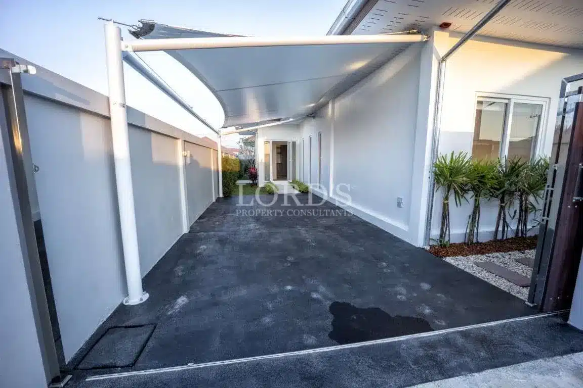 Covered driveway with modern shade canopy, smooth concrete flooring, white exterior walls, and landscaped plants beside a residential villa.