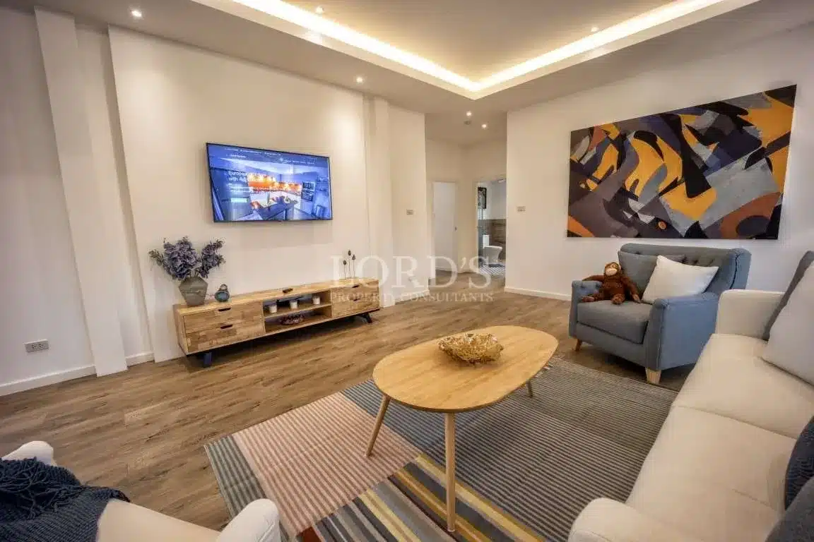 Modern living room with wall-mounted TV, wooden media console, neutral sofas, abstract wall art, and recessed ceiling lighting.