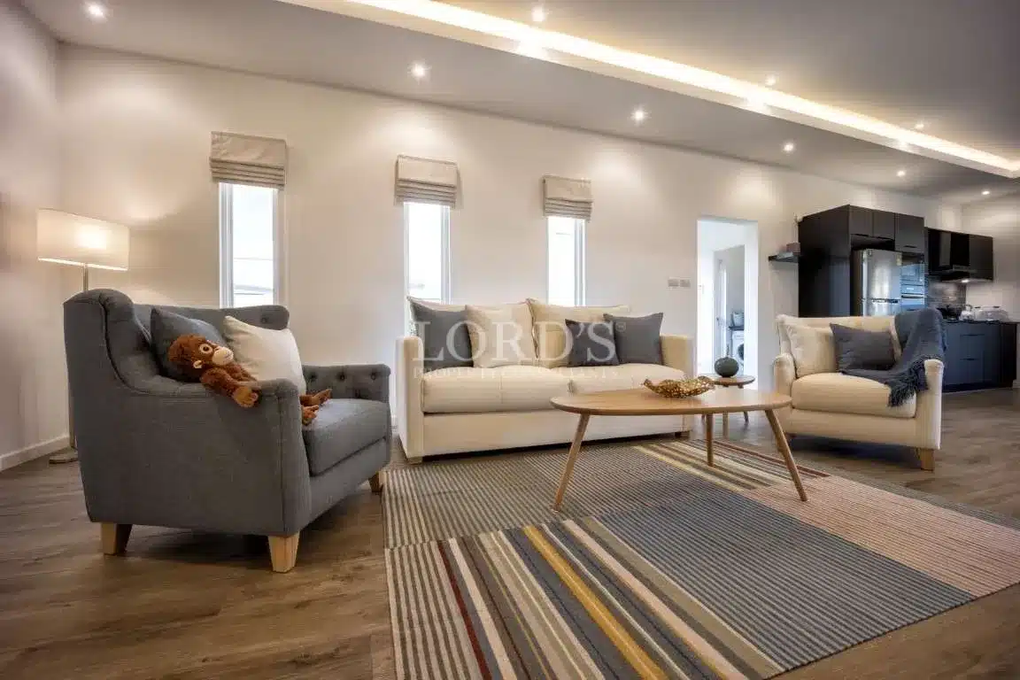 Modern open-plan living room with neutral sofas, armchair, wooden coffee table, striped area rug, and recessed ceiling lighting.