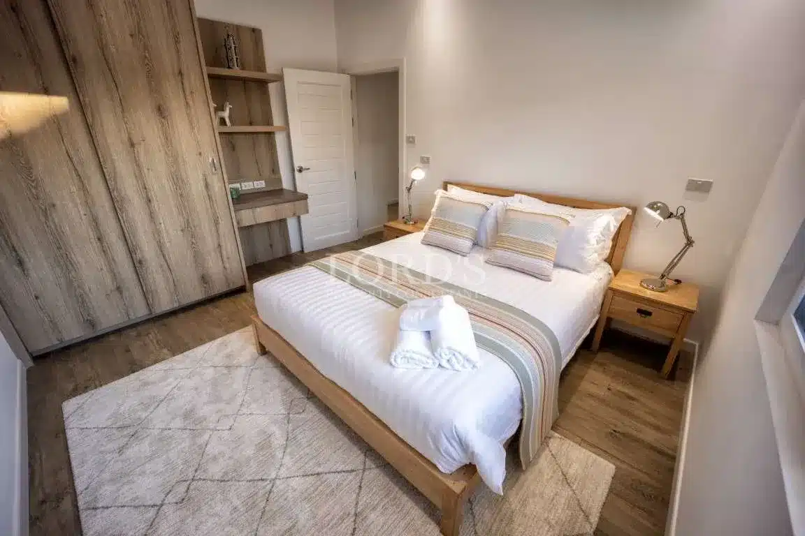 Modern minimalist bedroom with wooden bed frame, white bedding, built-in wardrobe, study desk, and warm ambient lighting.