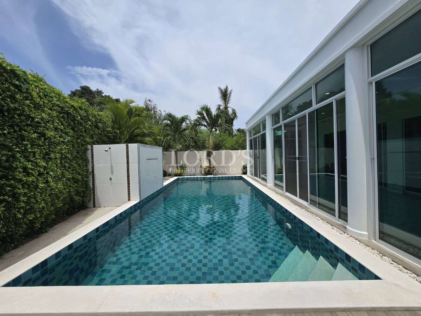 Private swimming pool with blue mosaic tiles beside a modern glass-walled villa surrounded by greenery.