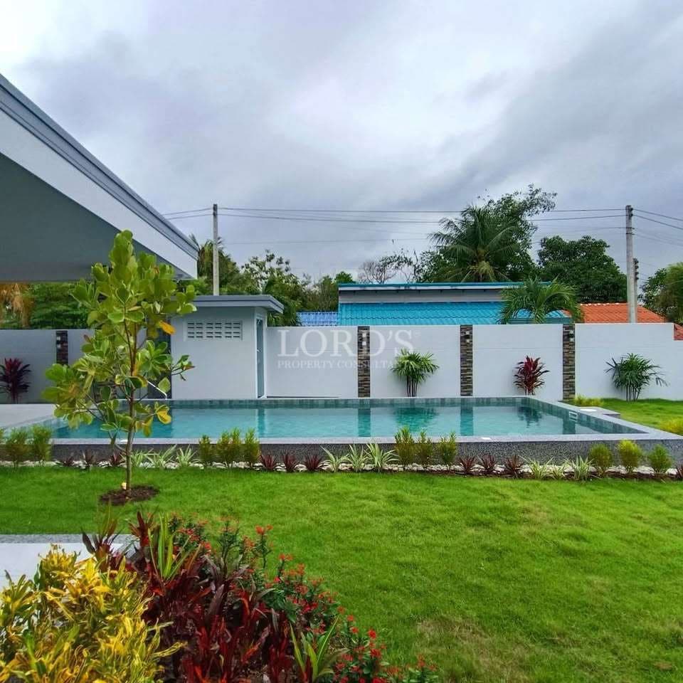 Private villa garden with rectangular swimming pool, manicured lawn, modern boundary wall, and tropical landscaping.
