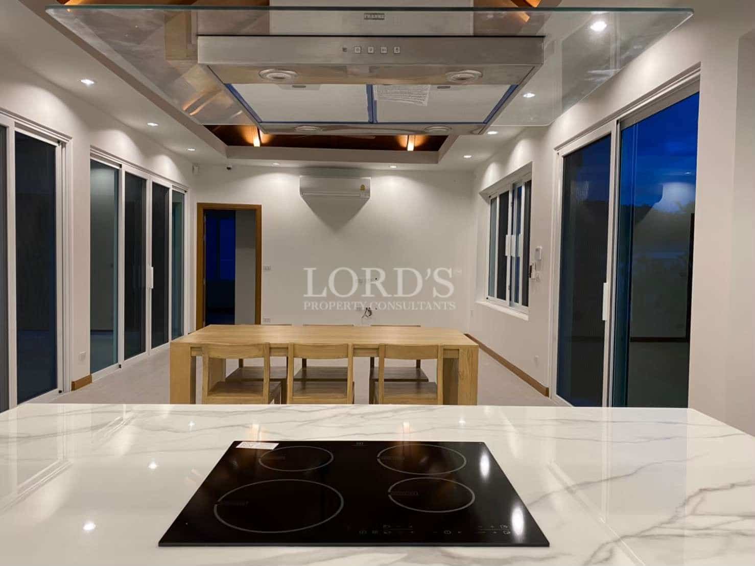 Modern open-plan kitchen and dining area with marble island, built-in cooktop, glass range hood, and large windows at night.