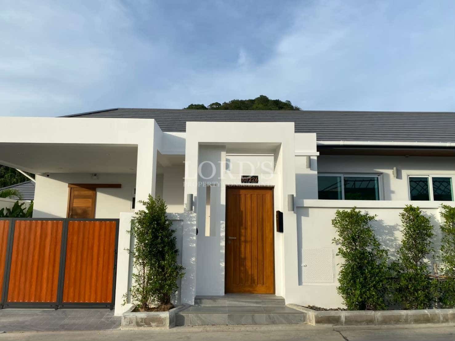 Modern villa exterior with minimalist white facade, wooden front door, private gate, and landscaped greenery.