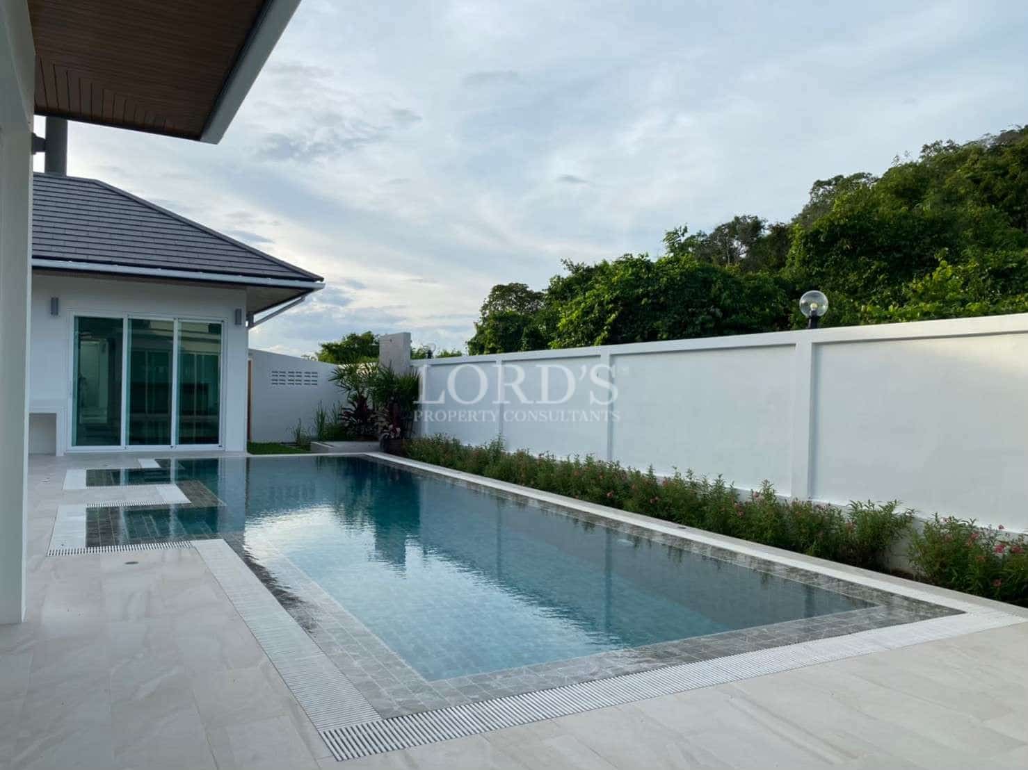 Private swimming pool beside a modern villa with tiled deck, white perimeter wall, and lush green surroundings.