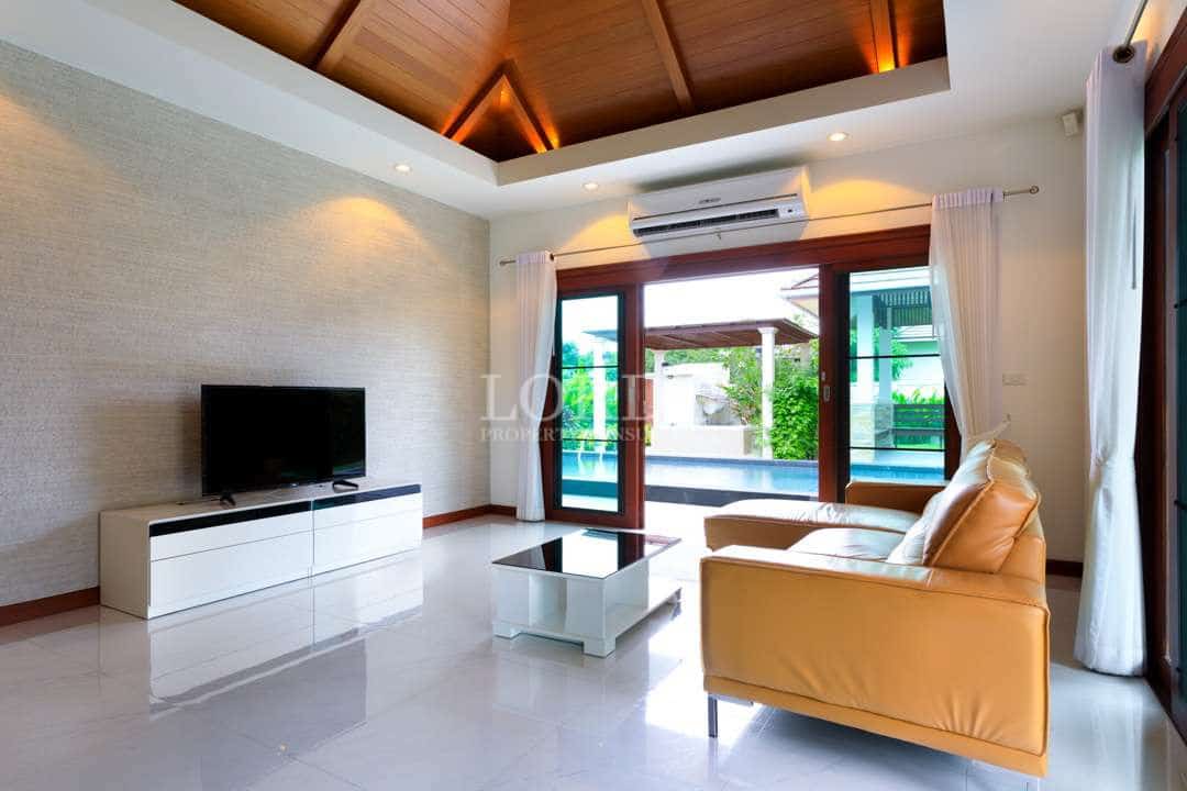 Luxury villa living room with high wooden ceiling, leather sofa, TV unit, and sliding glass doors opening to a private pool