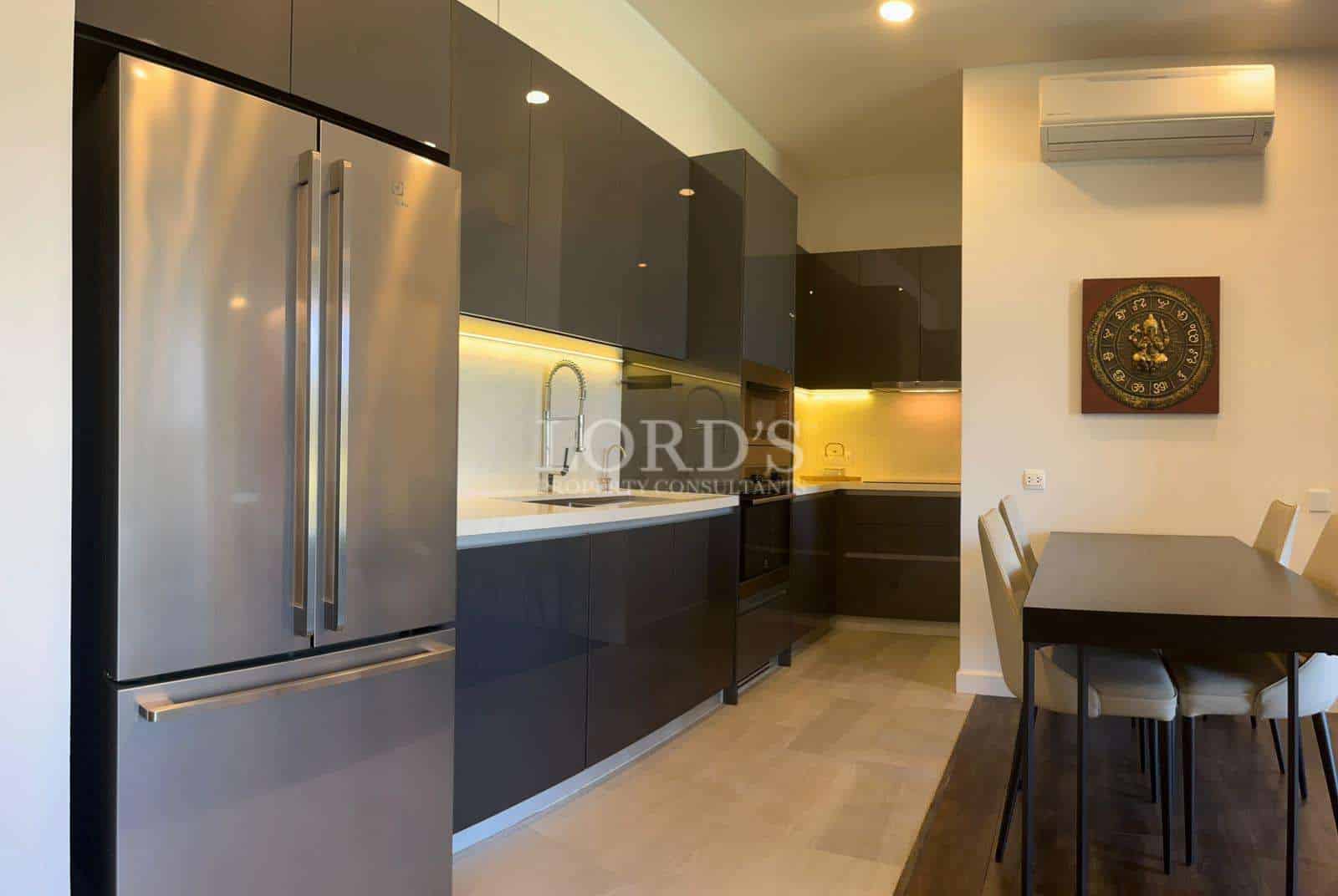 Modern open-plan kitchen with glossy dark cabinets, stainless steel refrigerator, built-in oven, warm under-cabinet lighting, and dining table in a contemporary apartment.